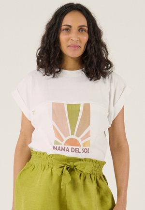 Vrouw met schouderlang krullend haar, gekleed in een wit T-shirt met een zonafbeelding en de tekst "MAMA DEL SOL", gecombineerd met groene hoge taillebroeken.