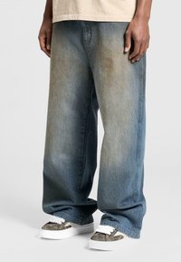 Baggy blå denimjeans med falmning og store frontlommer, parret med sneakers i camouflage mønster med hvide snørebånd og en hvid sål.