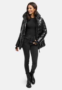 Sort pufferjakke med en blank finish, oversized hætte og elastiske manchetter. Bæret over en sort top og parret med mørke jeans og sorte støvler.
