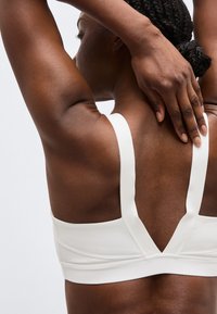 Γυναίκα με πλεξούδες, που φοράει λευκό αθλητικό σουτιέν, σηκώνει ένα χέρι και αγγίζει το πίσω μέρος του λαιμού της με το άλλο χέρι.