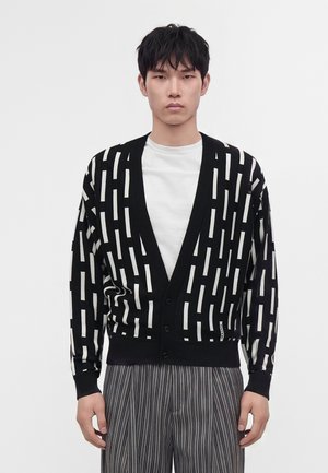 Jeune homme portant un cardigan noir avec des rayures verticales blanches, un t-shirt blanc et un pantalon gris à fines rayures, debout devant un fond uni.
