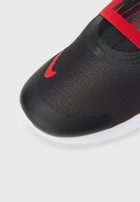 Primer plano de la punta de una zapatilla deportiva negra y roja con suela texturizada y parte superior de malla sobre fondo blanco.