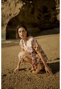 Kortärmad, knälång klänning i ett rosa och beige abstrakt blommönster, textilerad tyg; modell som hukar sig på en sandstrand, naturligt ljus.