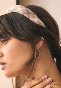 Beige ja ruskeasävyinen ruudullinen kangaspanta, yhdistettynä kierretyn muotoilun omaaviin kultaisiin renkorenkaihin. Hiukset laskeutuvat vapaasti sivulle.