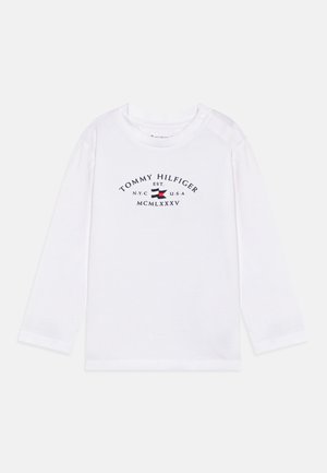T-shirt blanc à manches longues en coton, avec un logo brodé bleu marine comprenant du texte et un petit emblème rouge et bleu.