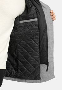 Chaqueta gris de mezcla de lana con un forro negro acolchado. Cuenta con cierre de cremallera y un bolsillo interior hecho de una tela gris en contraste.