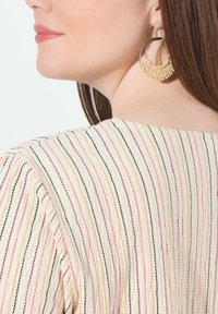 Beige gestreiftes Oberteil mit mehrfarbigen Linien in Pink, Orange und Schwarz; mit rundem Ausschnitt und strukturierter Stoffqualität. Große geflochtene Ohrstecker.
