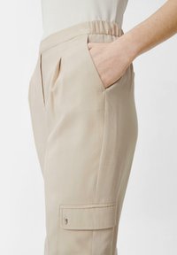 Beżowe spodnie cargo o gładkiej fakturze, elastycznym pasku, bocznych kieszeniach i jednej naszytej kieszeni na nogawce. Dopasowany krój z fałdami.