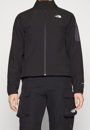 Person iført en sort North Face lynlåstrøje med høj krave og sorte cargobukser med store sidelommer.