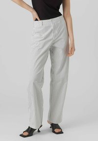 Pantalons rayés en blanc et noir, présentant une taille haute, une coupe droite et une texture de tissu lisse. Accompagnés de chaussures noires à bout ouvert.