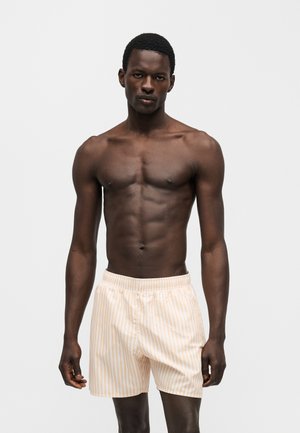 Modèle masculin à la peau foncée et aux cheveux courts, portant un short de bain clair à rayures, debout devant un fond blanc uni.