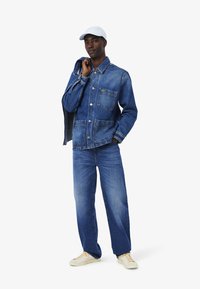 Giacca di denim con due tasche frontali, colore blu e chiusura a bottoni, abbinata a jeans coordinati e suole chiare. Il cappellino aggiunge un tocco casual.