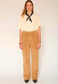 Haut beige à manches courtes avec un foulard noir à motifs, associé à un pantalon en velours côtelé taille haute et large en couleur beige chaud et des chaussures noires cloutées.