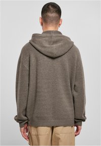 Brown knit hoodie with a relaxed fit, large hood, and ribbed hem. Soft texture and drop shoulder design; worn with light-colored pants.