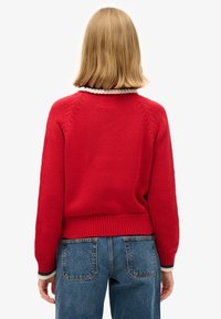 Rode gebreide trui met geribde zoom, voorzien van een gestreepte kraag. Gecombineerd met hoge blauwe spijkerbroeken. Textuur van het gebreide patroon is zichtbaar.