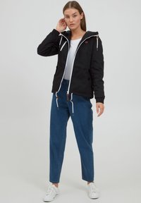 Jachetă neagră cu fermoar, șnururi albe, glugă și buzunare; asortată cu pantaloni din denim albastru și adidași albi. Material neted, design casual.