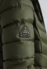 Veste matelassée vert olive avec texture en quilt. Présente un patch logo triangulaire noir sur l'épaule et une fermeture zippée.