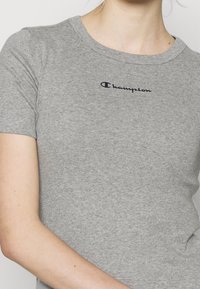 Šedé tričko s krátkým rukávem vyrobené z bavlny. Má kulatý výstřih a na přední straně vyšívané logo "Champion" v námořnické modři.