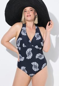 Námořnické jednodílné plavky s bílým spirálovým květinovým vzorem, s hlubokým výstřihem do V a zavazováním za krkem, doplněné širokým černým slaměným kloboukem.