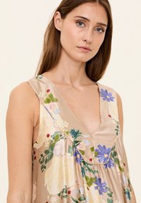 Ärmelloses floral Kleid in Beige mit tiefem V-Ausschnitt, das blaue und cremefarbene Blumen, grüne Blätter und geraffte Textur an der Brust zeigt.