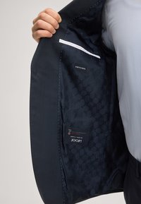 Wnętrze granatowego wełnianego żakietu, pokazujące wzorzystą podszewkę, metkę z napisem "Wool Stretch" oraz kieszeń z detalami brandingowymi.