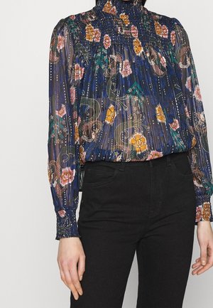 Sheer navy blue blouse with high neck, floral and paisley pattern in orange, pink, and green, paired with black high-waisted pants.