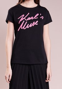 Černé bavlněné tričko s krátkým rukávem a kulatým výstřihem, který nese výrazný růžový grafický text "Karlova múza".