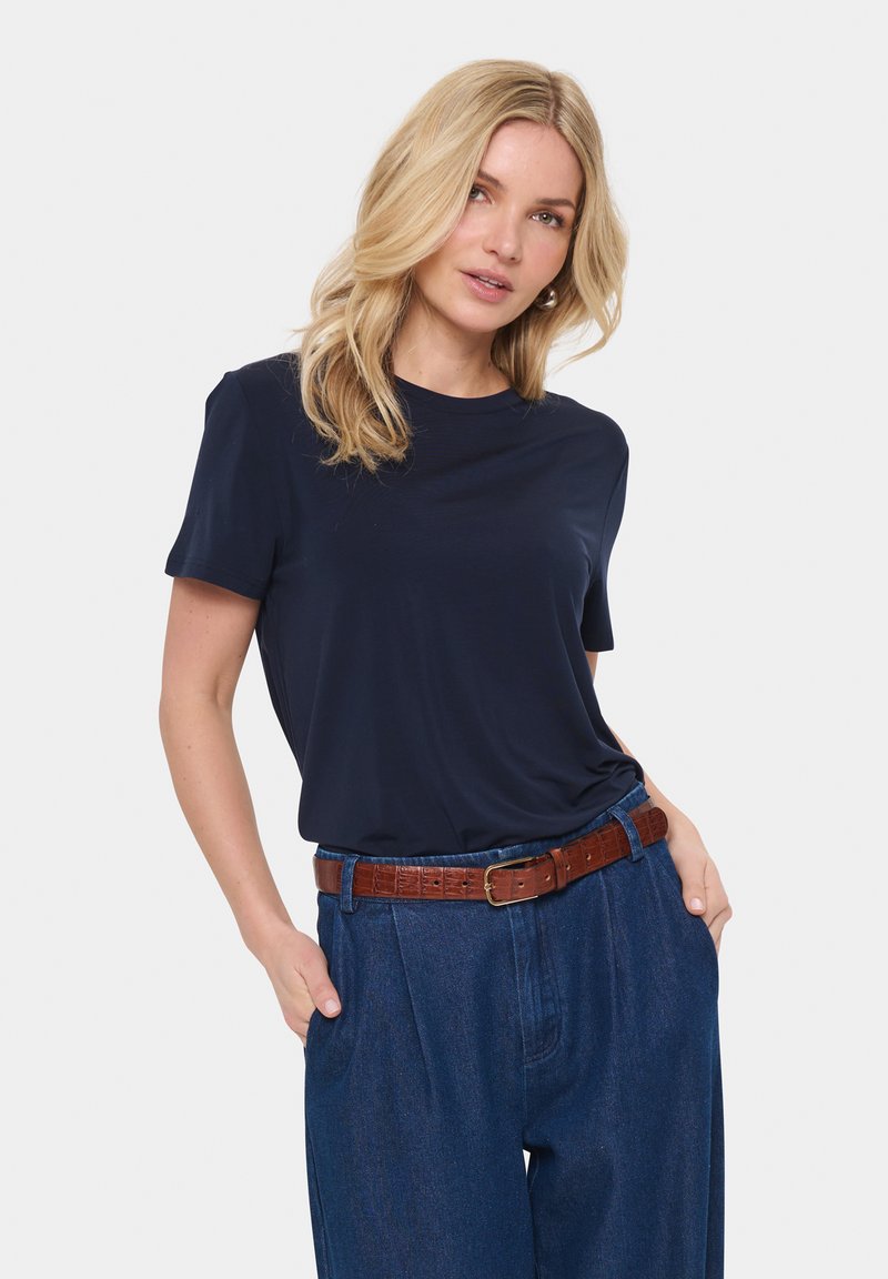 Sötétkék, rövid ujjú póló sima anyagból, magas derekú kék farmernadrággal és barna bőr övvel párosítva.