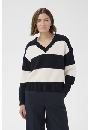 Femme aux cheveux bruns courts portant un pull à col en V rayé noir et blanc et un pantalon foncé, sur un fond clair uni.