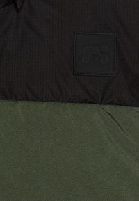 Augšpusē ir melna svītraina auduma daļa, bet apakšā ir vienkrāsaina tumši zaļa auduma daļa. Augšējā daļā ir teksturēts melns logotipa uzlīme.