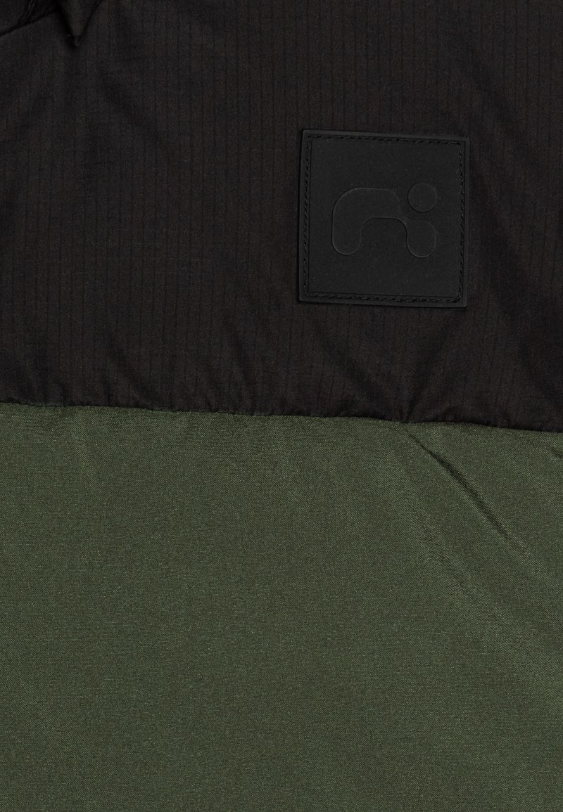 Zwart gestreepte stof aan de bovenkant, met daaronder een effen donkergroene stof. Bevat een gestructureerde zwarte logo-patch op het bovenste gedeelte.