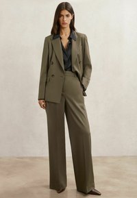 Donna in piedi davanti a uno sfondo semplice, indossa un blazer doppiopetto verde oliva, pantaloni a gamba larga abbinati e una camicia nera con bottoni.