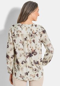 Blusa a maniche lunghe in tessuto leggero presenta una base beige chiaro con motivo floreale marrone e crema; scollatura arrotondata e leggero dettagli di piega sul retro.