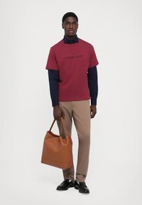 Bordeaux t-shirt med præget "MICHAEL KORS", lagdelt over en navy langærmet skjorte. Lysebrune bukser og en struktureret brun lædertaske.
