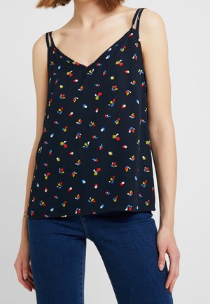 Woman wearing a black sleeveless V-neck top with red, blue, white, and yellow abstract shapes, paired with blue jeans.