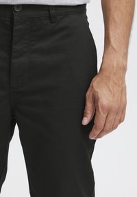 Black cotton trousers with a straight cut, featuring a button closure and side pockets. Close-up view of hand on the fabric.