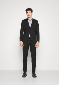 Giacca su misura nera con due bottoni, abbinata a una camicia lilla a fantasia, pantaloni neri e scarpe nere. Texture complessiva liscia.