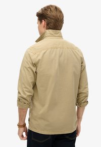 Veste beige avec un col plié, des poignets boutonnés et un ourlet droit. Fabriquée en tissu texturé, elle présente des accents de couture à l'arrière.