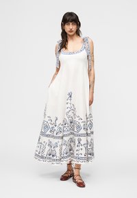 Γυναίκα που φοράει μακρύ λευκό φόρεμα με μπλε λουλουδένια και γεωμετρικά μοτίβα, με δεμένους ιμάντες στους ώμους και καφέ δερμάτινα παπούτσια με κορδόνια, που στέκεται μπροστά σε απλό φόντο.