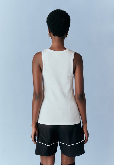 Débardeur blanc à côtes avec des bretelles larges, associé à un short noir avec piping blanc, vu de dos sur un fond bleu clair.