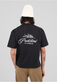 Czarny t-shirt z krótkim rękawem z białym napisem "Zabronione" oraz grafiką liścia na plecach. Noszony z beżowymi spodniami i białą wełnianą czapką.