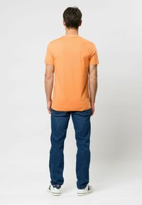 Tricou scurt cu mânecă scurtă, de culoare portocalie, cu guler rotund, purtat împreună cu o pereche de blugi albaștri și adidași albi, văzut din spate. Design simplu și lejer.