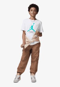 Hvit T-skjorte med en turkise Jordan-logo, beige genser knyttet rundt livet, brune cargo-bukser og grå sneakers med hvite detaljer.