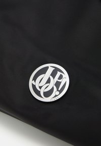 Czarna torba z materiału z wyraźnym, okrągłym srebrnym logo z stylizowanymi literami i napisem "JEANS" u dołu. Teksturalna powierzchnia.