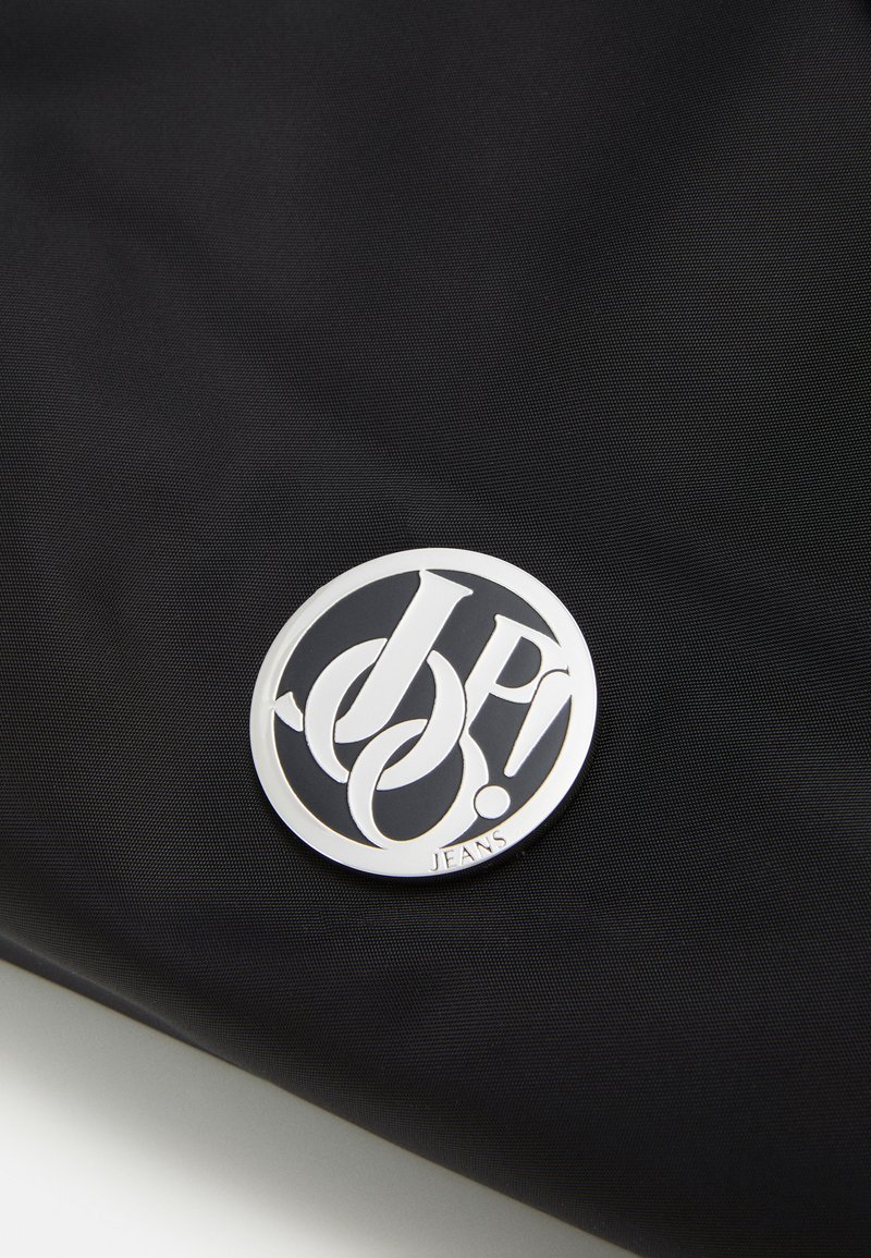 Schwarze Stofftasche mit einem auffälligen runden silbernen Logo, das stilisierte Buchstaben und das Wort "JEANS" am Boden zeigt. Strukturierte Oberfläche.