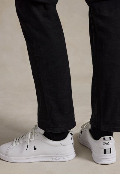 Weiße Polo Ralph Lauren Sneaker, getragen mit schwarzen Hosen und schwarzen Socken, von Seiten- und Rückansicht auf grauem Boden gezeigt.