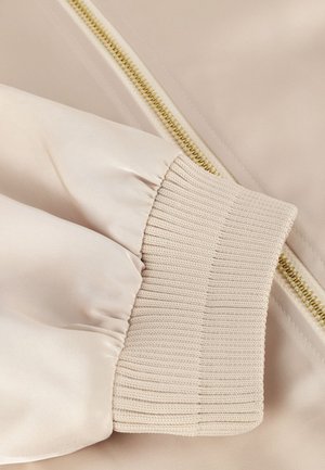 Beige bomberjack van een stof met een glanzend oppervlak. Voorzien van een ribbels manchet en een contrasterende gouden rits aan de zijkant, die de soepele textuur benadrukt.
