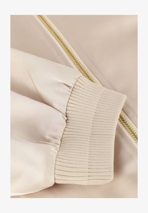 Beige bomberjack van een stof met een glanzend oppervlak. Voorzien van een ribbels manchet en een contrasterende gouden rits aan de zijkant, die de soepele textuur benadrukt.