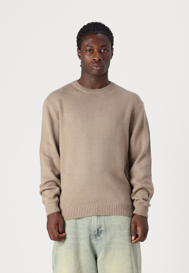 Beiger Strickpullover mit Rundhalsausschnitt und gerippten Bündchen, aus einem strukturierten Stoff und in lässigem Schnitt, kombiniert mit hellblauen Jeans.