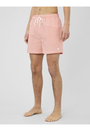 Homme portant un short de bain rayé rose et blanc jusqu'aux genoux avec cordon de serrage, debout pieds nus sur un fond gris.
